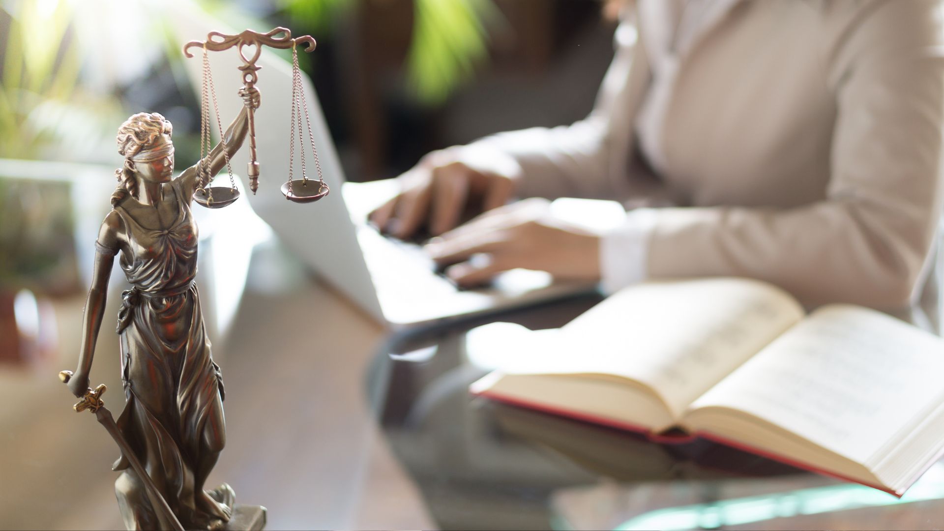 Blind Lady Jusitice statue in front of a blurred professional on a laptop with an open book to represent how the Rosemont College Professional Studies Criminal Justice BS degree is part of why Rosemont College is a Forbes Rated best online college and a best Main Line college.