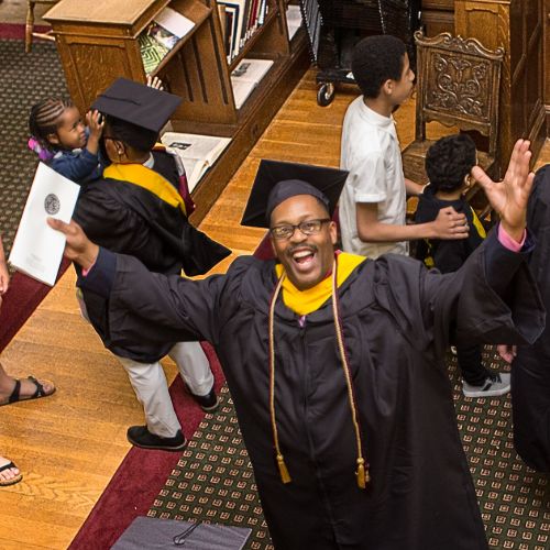 Smiling graduate holding his diploma with his hands up in celebration to represent how Rosemont College alumni are part of why Rosemont College is a Forbes rated best online college, and a best mainline college.