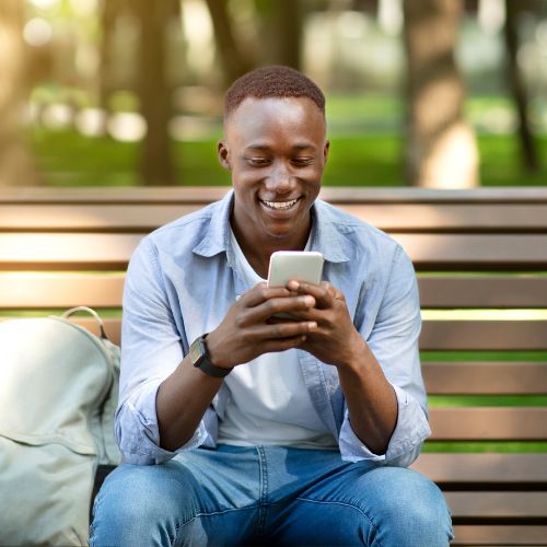 African American man seated on a bench smiling and using the Rosemont app on his phone to represent how technology is part of why Rosemont College is a Forbes rated best online college, and a best Main Line college.