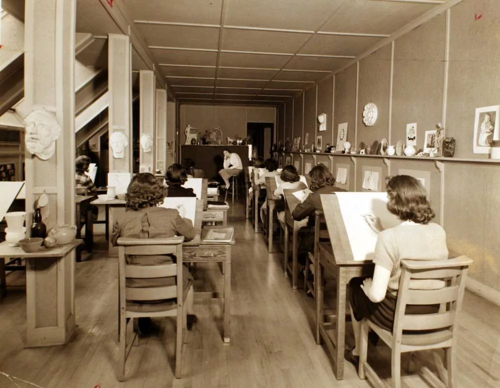 Image of a Rosemont College art classroom in the 1930's. The back of all the female student's heads are visible as they draw a clothed model in a knee-length skirt leaning over on a stool with her elbow on her knee. This image is to help represent how the history of art classes is part of why Rosemont College is a Forbes rated best online college, and a best Main Line college.