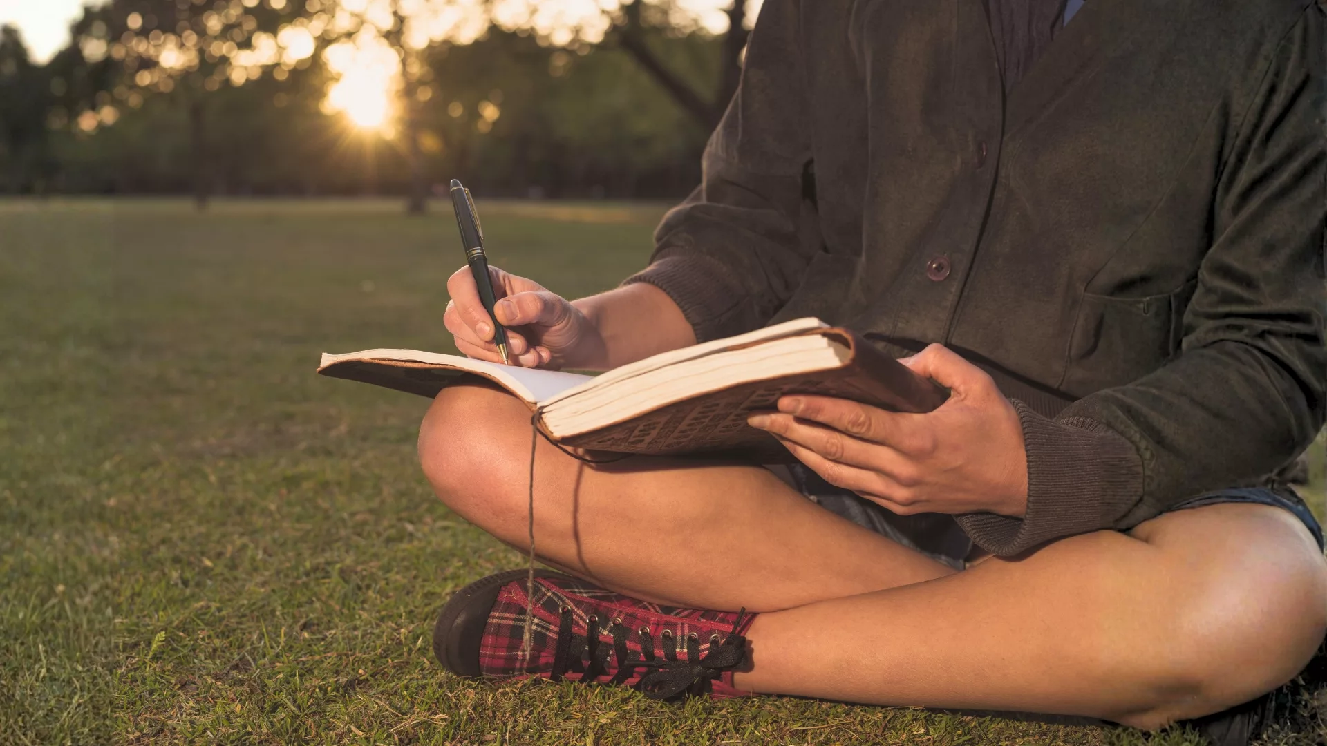 Rosemont College Writer's Studio - Memoir As Literary Nonfiction Masterclass Close up of a woman sitting on grass at sunset writing in a journal to represent how the Writer's Studio Literary Nonfiction Masterclass is part of why Rosemont College is a Forbes rated best online college, and a best Main Line college.